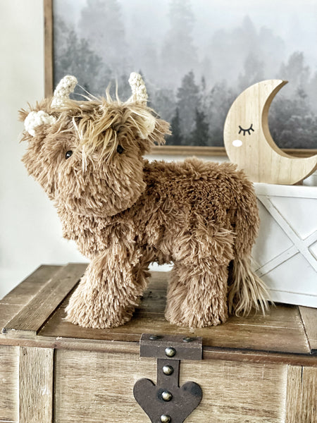 A plush standing Highland Cow toy with a brown coat, positioned on a wooden surface with a decorative moon-shaped item in the background.