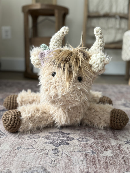 A plush highland cow stuffed animal with tan faux fur. This highland cow is handmade by ClaraLoo with crocheted accents.
