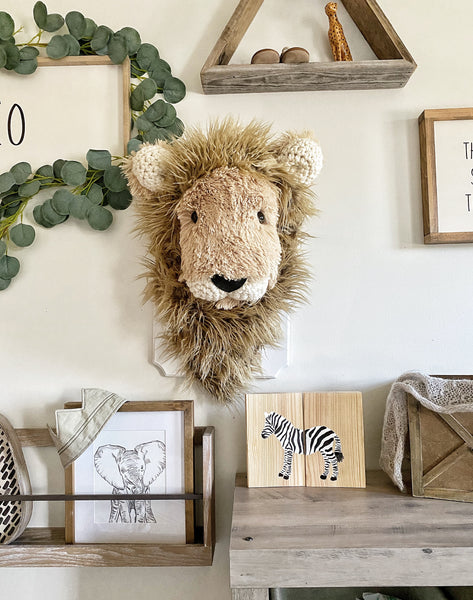 A plush lion head made by ClaraLoo that is mounted on a white wooden plaque in a safari themed nursery. The stuffed lion has soft tan fur, a shaggy brown mane, and crocheted accents.